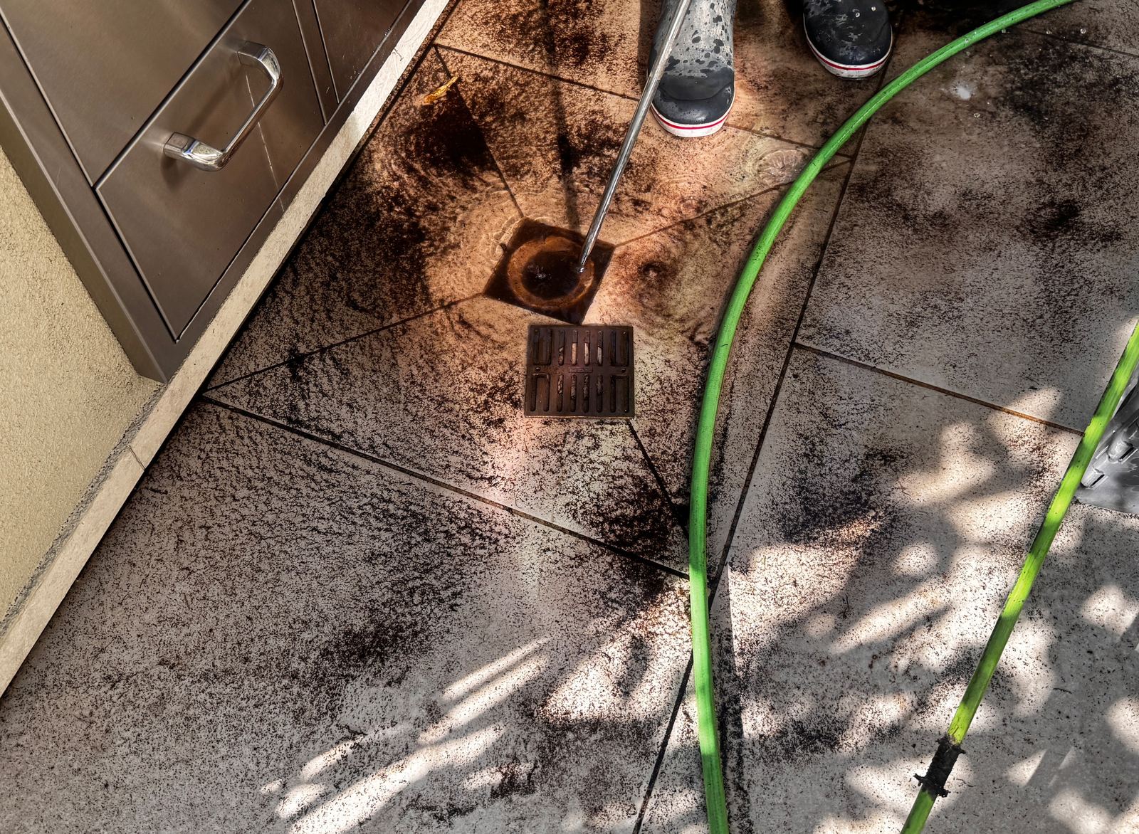 Patio drain beside a stainless outdoor kitchen being flushed, with dark debris spread across the tile
